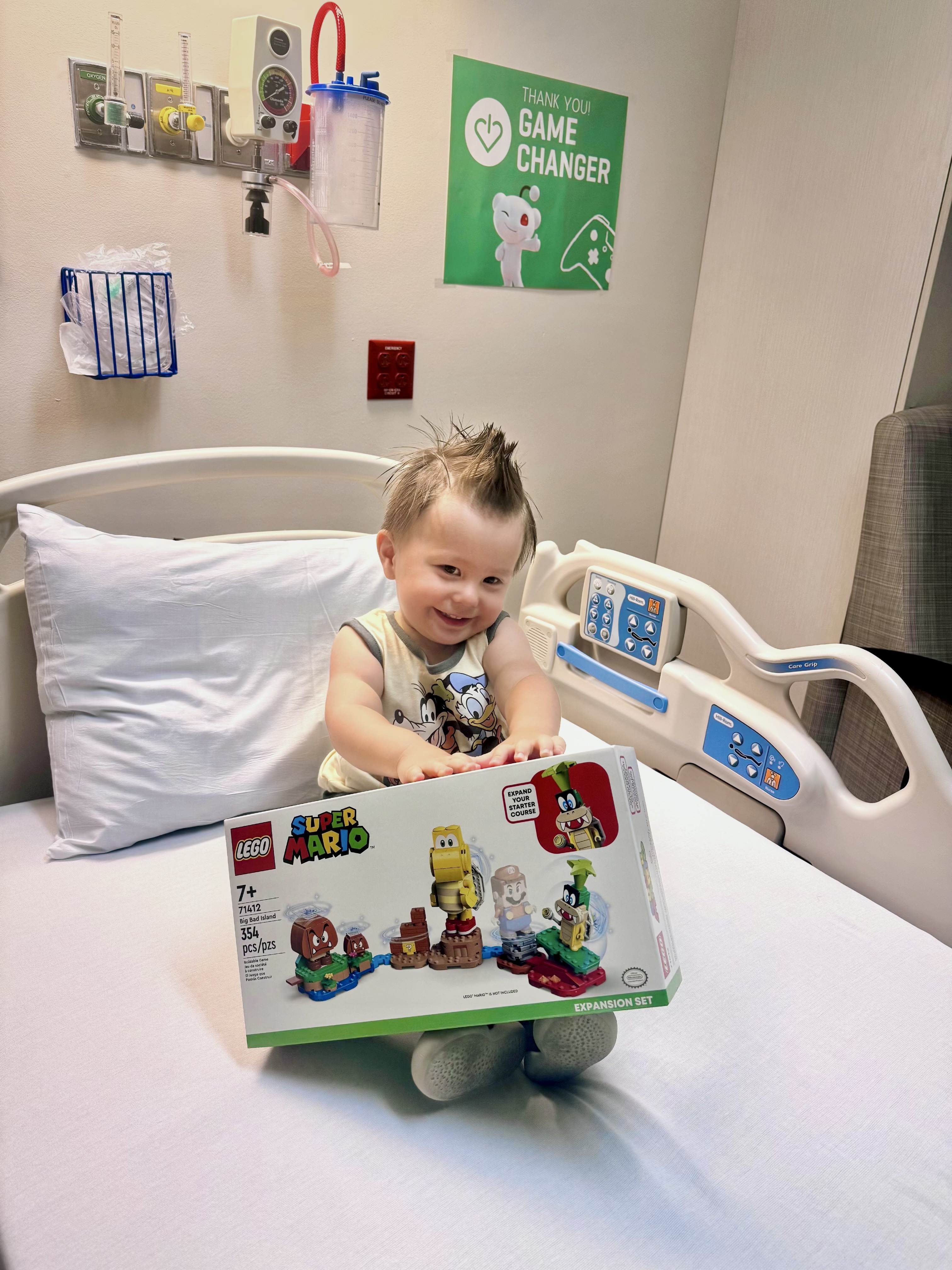A smiling toddler in a hospital bed with a LEGO gift and a Thank You GameChanger poster.